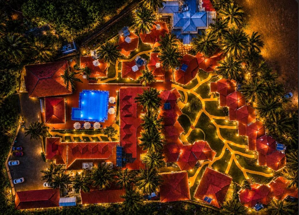 A property seen from directly above at night, lit pathways winding between red-tiled roofs, palms and a floodlit pool