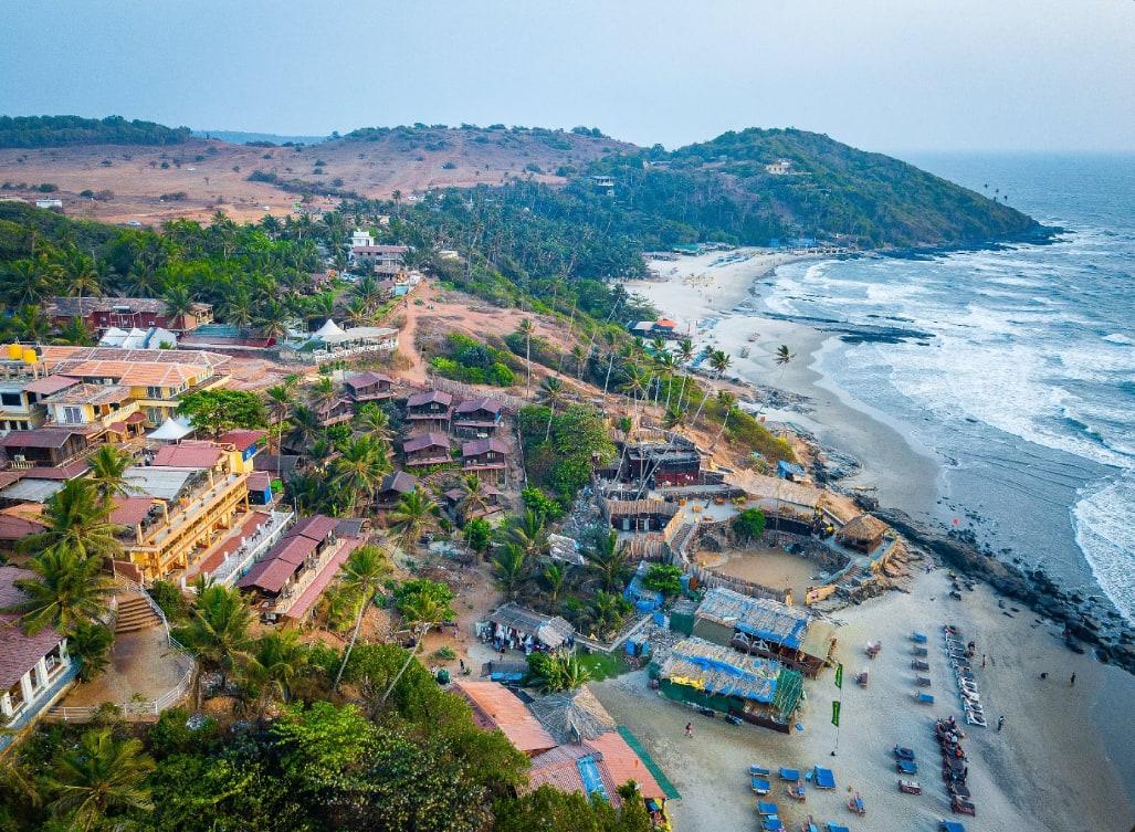 An aerial view of a headland settlement above a beach, huts along the slope and surf breaking on the rocks