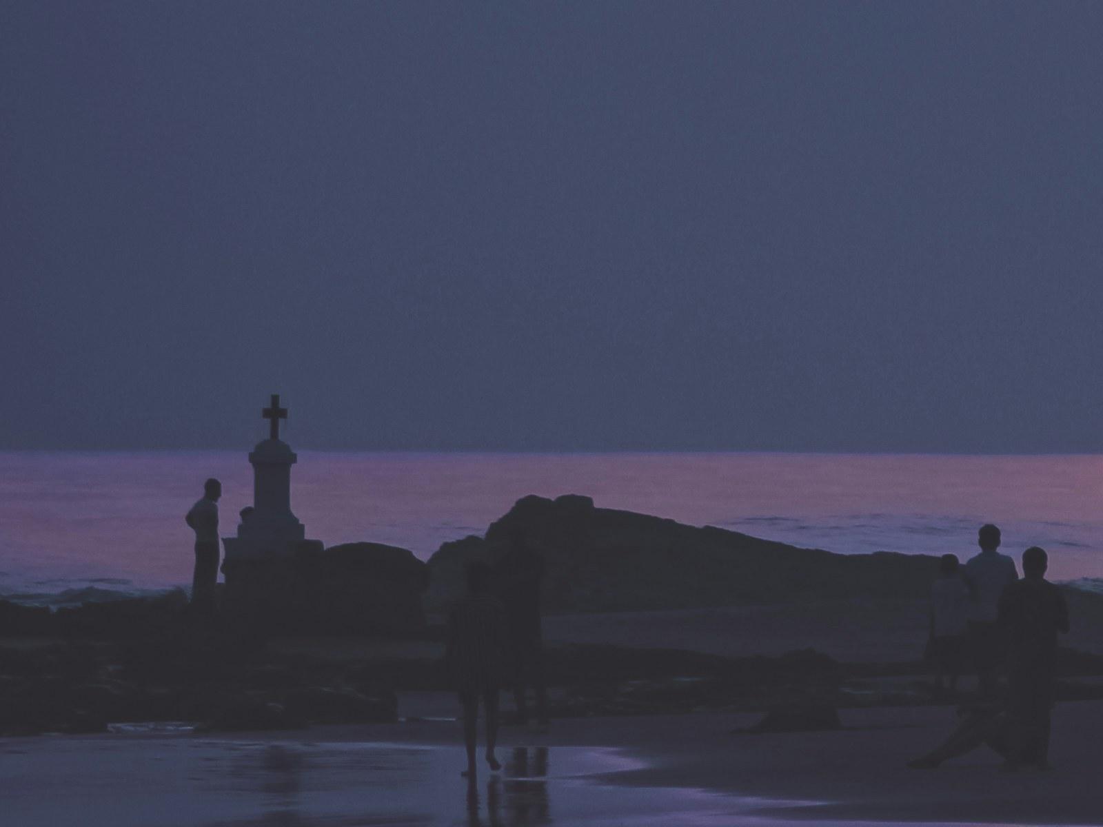 A beach at dusk with figures silhouetted along the waterline, and a small stone cross on a stepped plinth standing among the rocks at the left