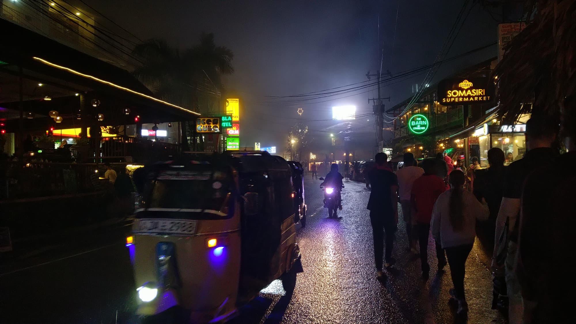Ella's main street at night: neon bar signs, a tuk-tuk, people out walking