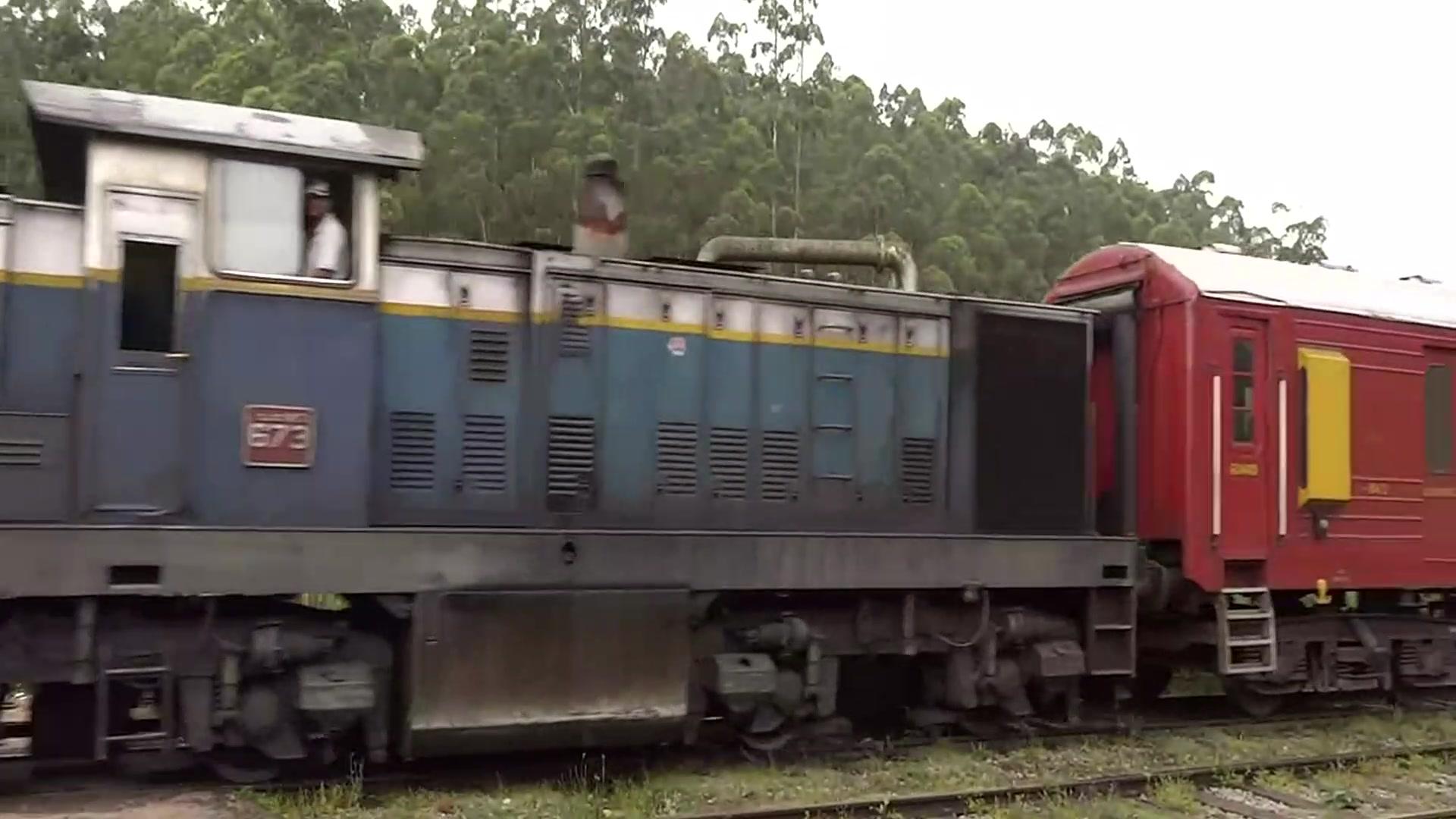 A diesel train with red carriages pulling through a hill-country station