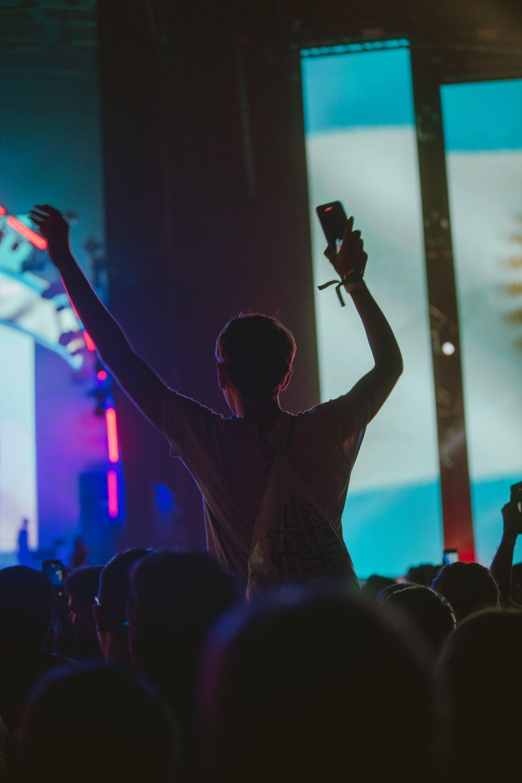 A figure with both arms raised in a festival crowd, magenta stage light behind